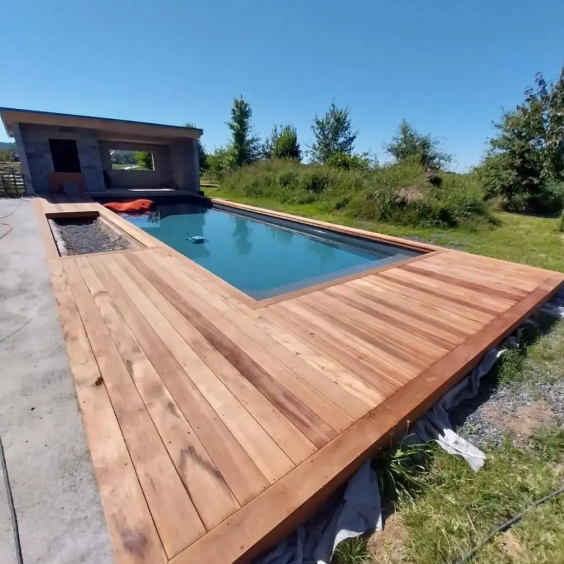 Terrasse en bois entourant une piscine moderne avec un pool house en pierre, sous un ciel bleu et entourée de verdure