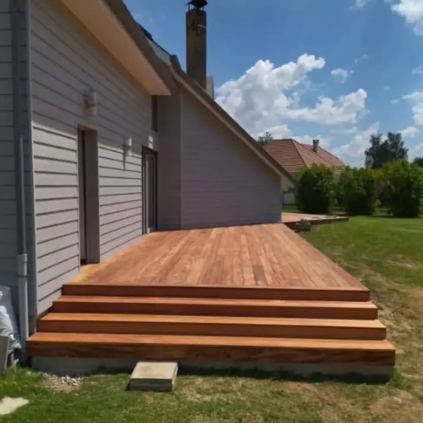 Terrasse en bois avec escalier intégré, attenante à une maison moderne en bardage gris, donnant sur un jardin verdoyant sous un ciel bleu.