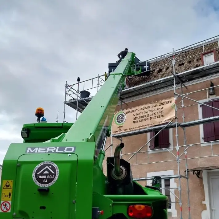 Travaux de rénovation de toiture sur une maison en pierre, avec un ouvrier sur un échafaudage et un chariot télescopique vert de marque Merlo soulevant du matériel