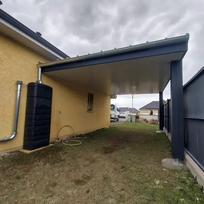 Aménagement extérieur d'une maison avec un carport moderne en métal et une cuve de récupération d'eau de pluie, offrant une protection pour véhicules et une solution écologique.