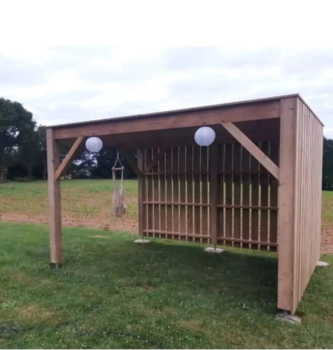Abri de jardin en bois avec toit plat, installé sur une pelouse, équipé de lanternes décoratives et d'une chaise suspendue pour un aménagement extérieur chaleureux.