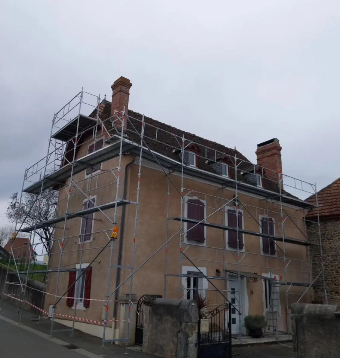 Maison ancienne entourée d'un échafaudage pour des travaux de rénovation de toiture et de façade, avec des cheminées en briques et des volets rouges.