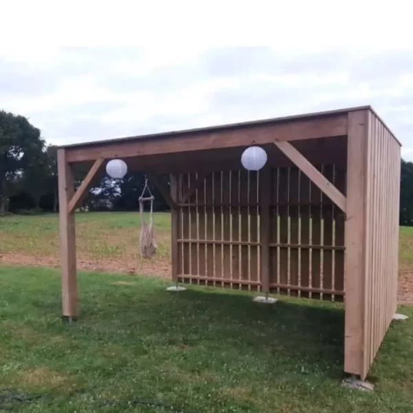 Abri de jardin en bois avec toit plat, installé sur une pelouse, équipé de lanternes décoratives et d'une chaise suspendue pour un aménagement extérieur chaleureux.