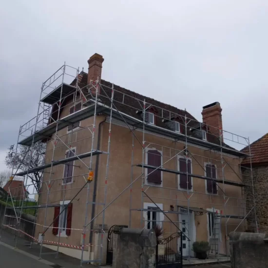 Maison ancienne entourée d'un échafaudage pour des travaux de rénovation de toiture et de façade, avec des cheminées en briques et des volets rouges.